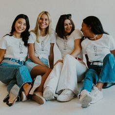 four women are sitting on the floor and smiling together, all wearing white t - shirts