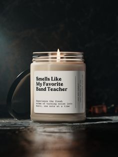 a candle sitting on top of a wooden table next to a black mug with the words smell like my favorite band teacher