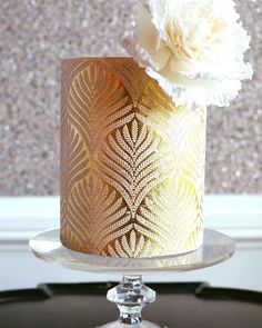 there is a gold and white decorated cake on a glass stand with a flower in it