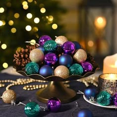 a table topped with a cake plate covered in purple and green christmas ornaments next to a lit candle