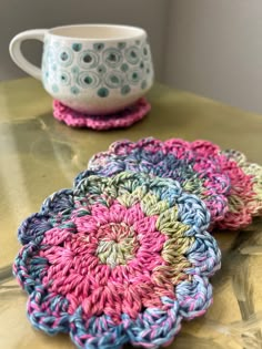 two crocheted coasters sitting on top of a wooden table next to a cup