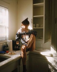 a woman sitting on top of a kitchen counter next to a sink and cupboards