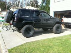 a black jeep parked in front of a house with large tires on it's back