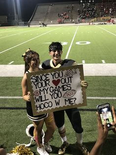 two girls holding up a sign on the sidelines at a football game, one girl has her cell phone in front of her