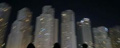 two people walking in front of tall buildings at night with the city lights shining on them