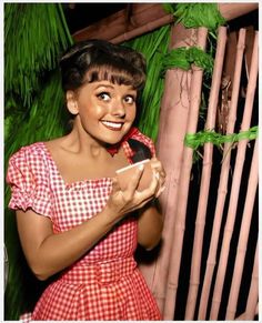 a woman in a red and white checkered dress holding a cell phone with palm trees behind her