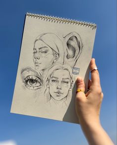 a hand holding up a drawing of three women's faces with the sky in the background