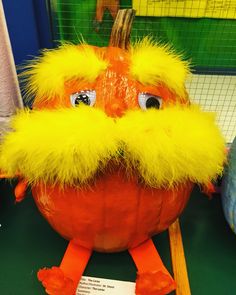 an orange pumpkin with yellow feathers on it's head and legs, sitting on a green surface