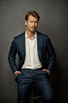 a man sitting on top of a stool wearing a blue suit and white shirt with his hands in his pockets