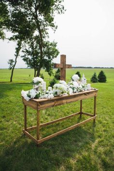 a wooden table topped with white flowers next to a cross in the middle of a field