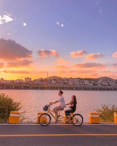 a man and woman riding on a bike next to the water at sunset or sunrise