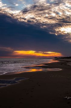 the sun is setting over the ocean with clouds in the sky and footprints on the sand