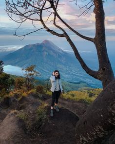 a woman standing on top of a dirt hill next to a tree and a mountain