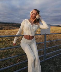 a woman leaning on a fence with her headphones in her ears and wearing white clothing