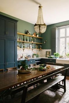 a kitchen with green walls and wooden table surrounded by shelves filled with potted plants