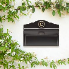 a black mailbox sitting on top of a white wall next to green leaves and flowers