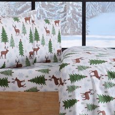 a bed covered in christmas themed sheets and pillows next to a window with snowy trees