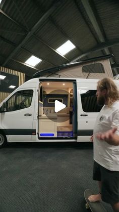 a man standing next to a white van in a garage with the door open and his skateboard on the ground