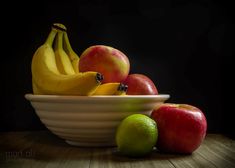 apples, bananas and oranges in a white bowl on a wooden table against a black background