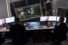 two men sitting at a desk with multiple monitors on it and one man standing in front of them