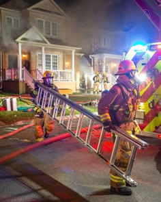 two fire fighters standing next to a ladder in front of a house at night with the lights on