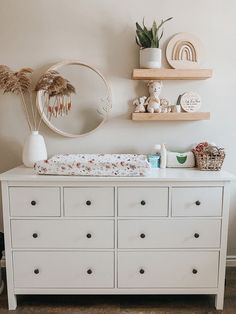 a white dresser topped with lots of drawers next to a wall mounted mirror and potted plants