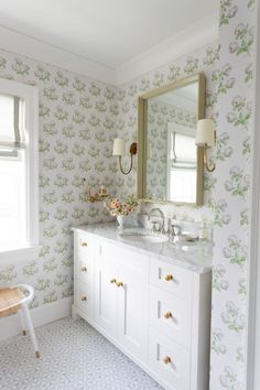 a white bathroom with wallpaper and a large mirror above the sink, along with a stool