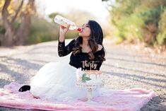 a woman sitting on a pink blanket drinking from a bottle while wearing a black shirt and white skirt