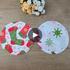 two christmas themed coasters sitting on top of a wooden table next to flowers and a potted plant