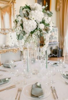 there is a table set with silverware and white flowers in a tall glass vase