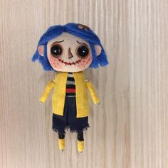 a small doll with blue hair and black eyes on a wooden surface next to a white wall