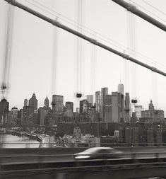 a black and white photo of a car passing by a bridge with the city skyline in the background