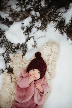 a baby is laying in the snow wearing a pink coat and a brown knitted hat