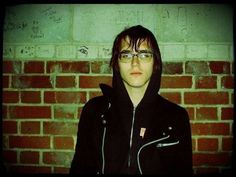 a woman with glasses standing in front of a brick wall wearing a black jacket and hoodie