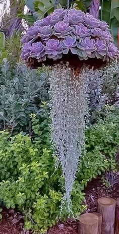 an umbrella shaped fountain in the middle of a garden with purple flowers and greenery