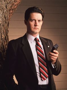 a man wearing a suit and tie holding a cell phone in his hand while standing next to a tree