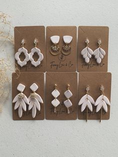 six pairs of earrings are displayed on brown cards with white flowers in the middle and gold accents