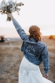 a woman in a white dress and jean jacket holds flowers up to the sky with her hand