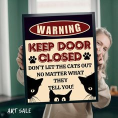 a woman holding up a sign that says warning keep door closed don't let the cats out no matter what they tell you