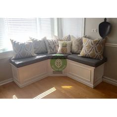 a corner bench with many pillows on it in front of a window and light coming through the blinds