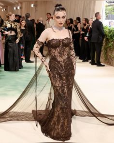 a woman in a brown dress standing on a white carpet with people behind her wearing black and gold