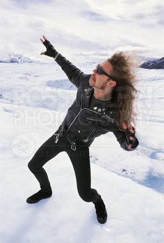 a man with long hair and sunglasses standing in the snow, arms outstretched to the sky