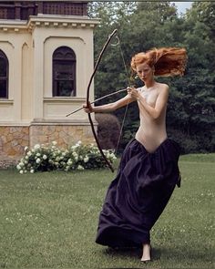 a naked woman holding a bow and arrow in front of a large building with trees