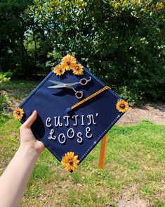 a person holding up a graduation cap with scissors and daisies on it, in front of some trees