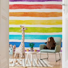 a giraffe standing in front of a rainbow painted wall with two stuffed animals