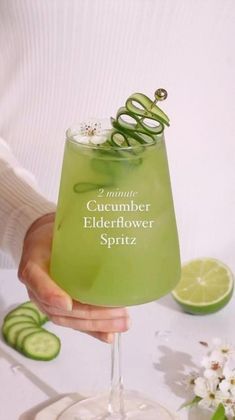 a person holding a green drink with cucumber on the rim and in front of it