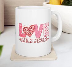 a white coffee mug with the words love like jesus on it next to a bowl of broccoli