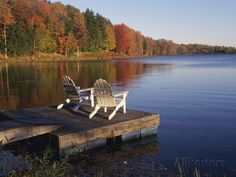 two chairs sitting on a dock in front of a body of water surrounded by trees