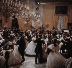 a group of people dressed in formal wear dance around a chandelier filled room