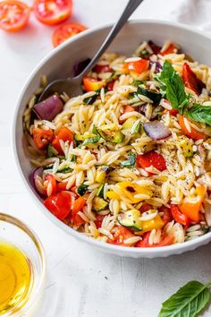 a white bowl filled with pasta salad next to a glass of olive oil and tomatoes